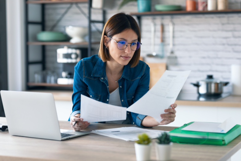 Shot of pretty young business woman working with computer while consulting some invoices and documents in the kitchen at home.